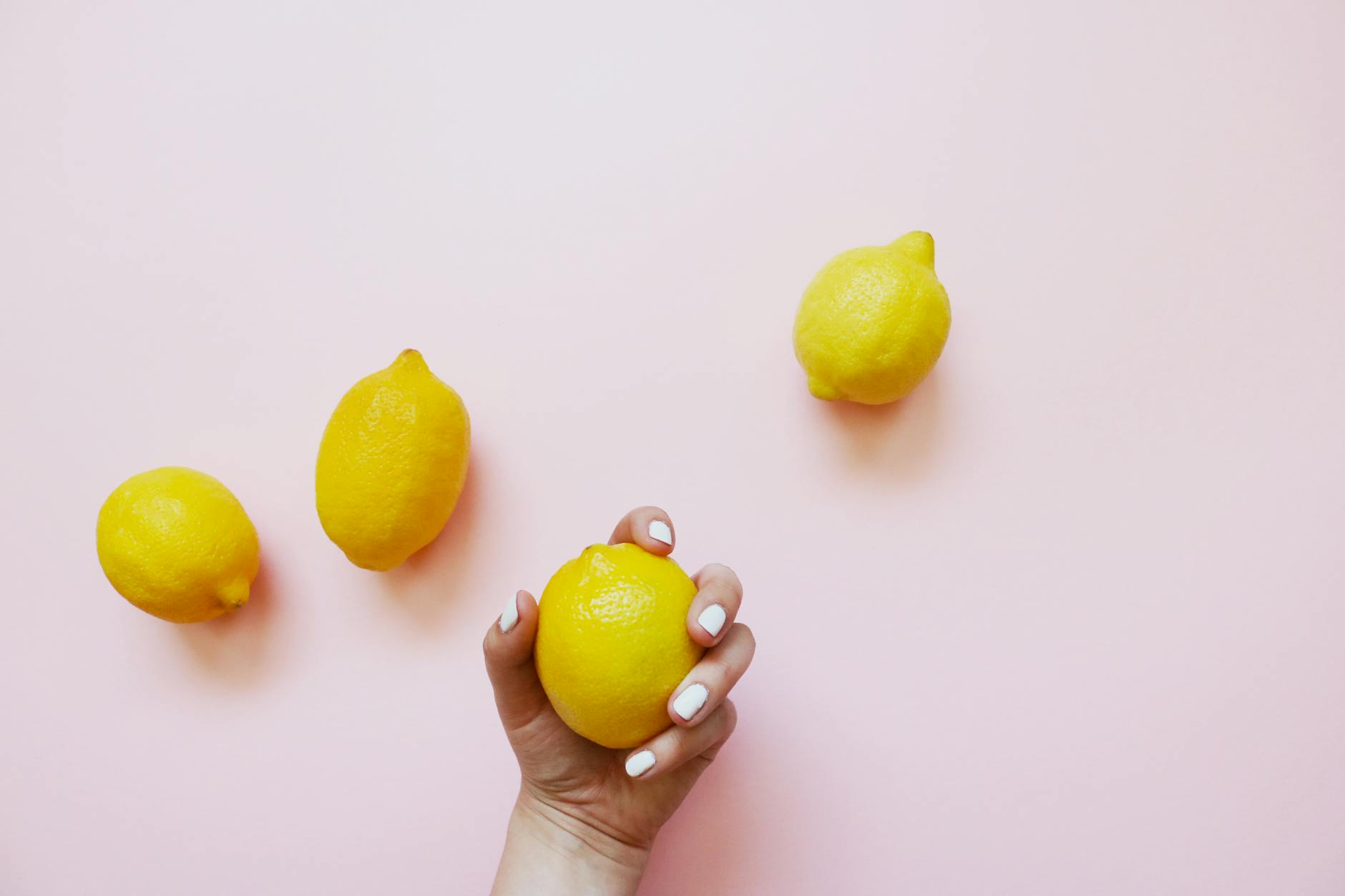 A hand holding a lemon on a soft pink background surrounded by additional lemons, symbolizing the citrus-inspired design of clitoral vibrators for pleasure.