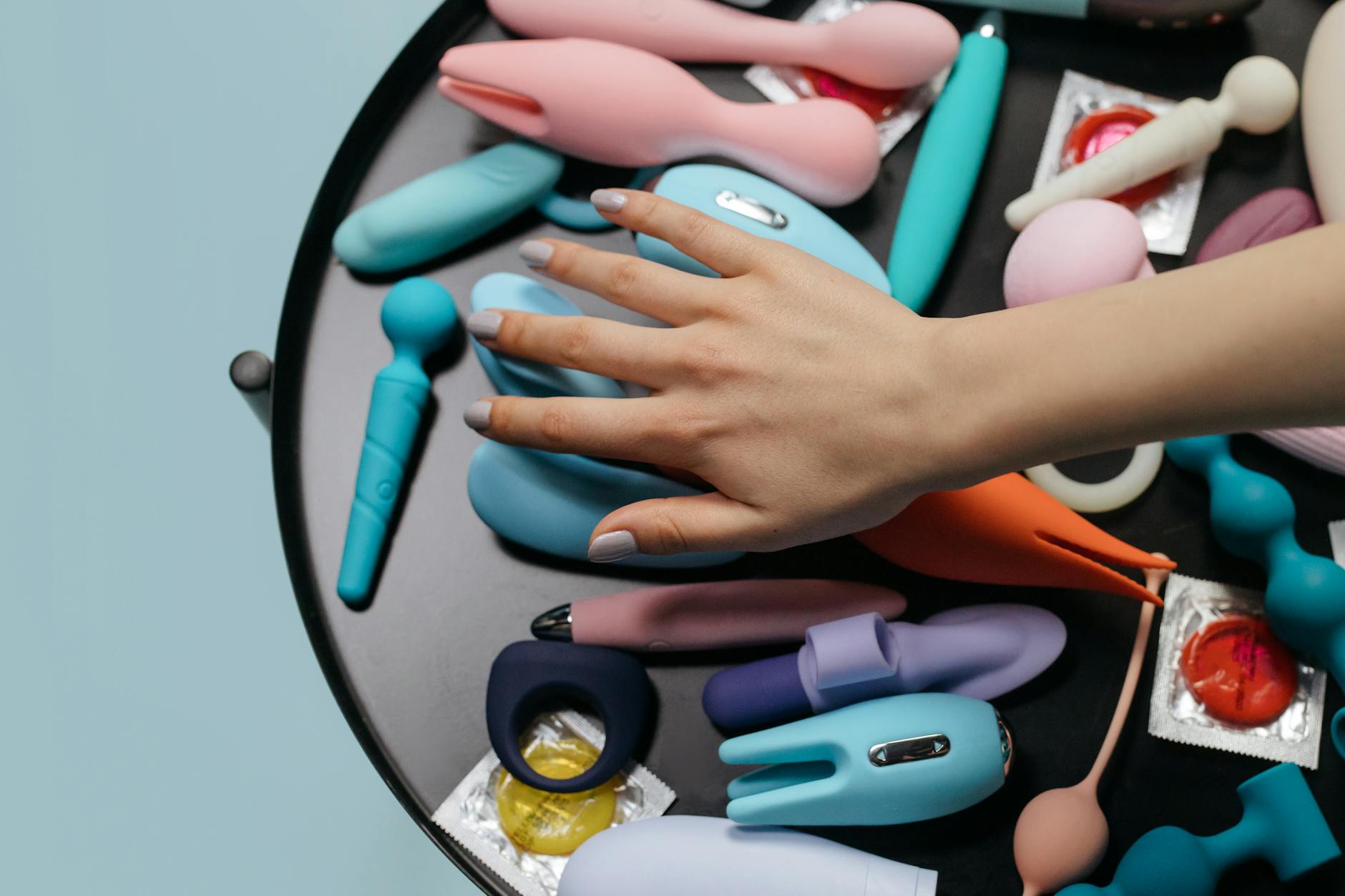 A hand selecting from a variety of colorful vibrators and intimate wellness tools on a soft surface.