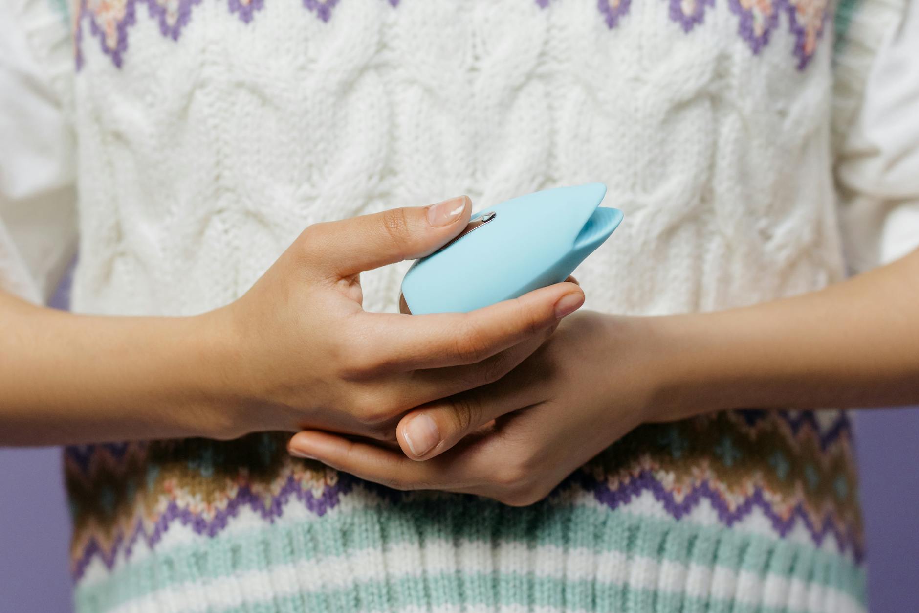 Hands holding a blue clitoral vibrator against a knitted sweater, representing intimate self-care